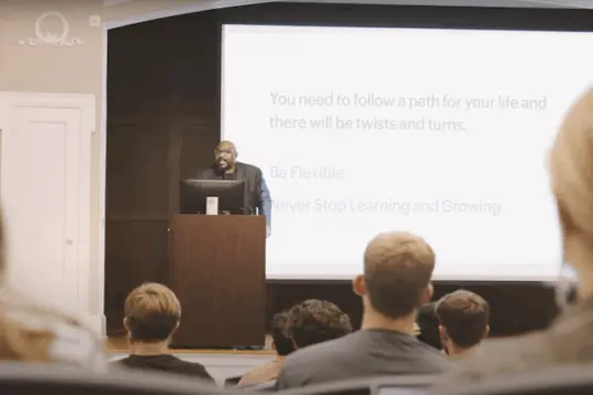 A speaker stands at a podium in front of a classroom, addressing students. Behind him is a large screen displaying a slide with the text: "Be flexible. Never stop learning and growing.